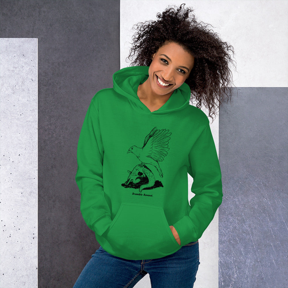 Irish green colored unisex Reflections hoodie. Features image of a crow with wings outstretched sitting on a sheep skull.  Has a front pouch pocket and double lined hood. Shown on female model against grey and white wall.