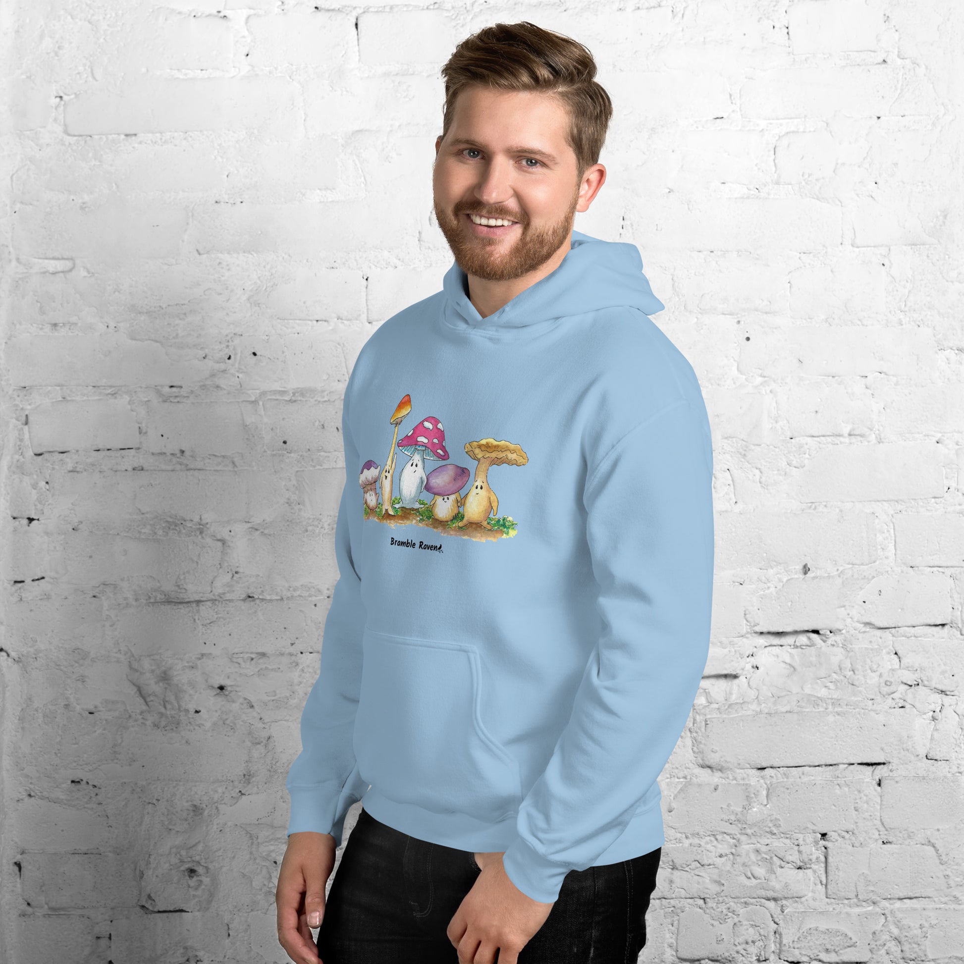 Light blue colored unisex cotton/polyester blend hoodie. Features front design of Mushy and his whimsical mushroom friends. Hoodie has double lined hood, front pouch pocket, rib knit cuffs and stretchy waistband. Shown on male model facing left.