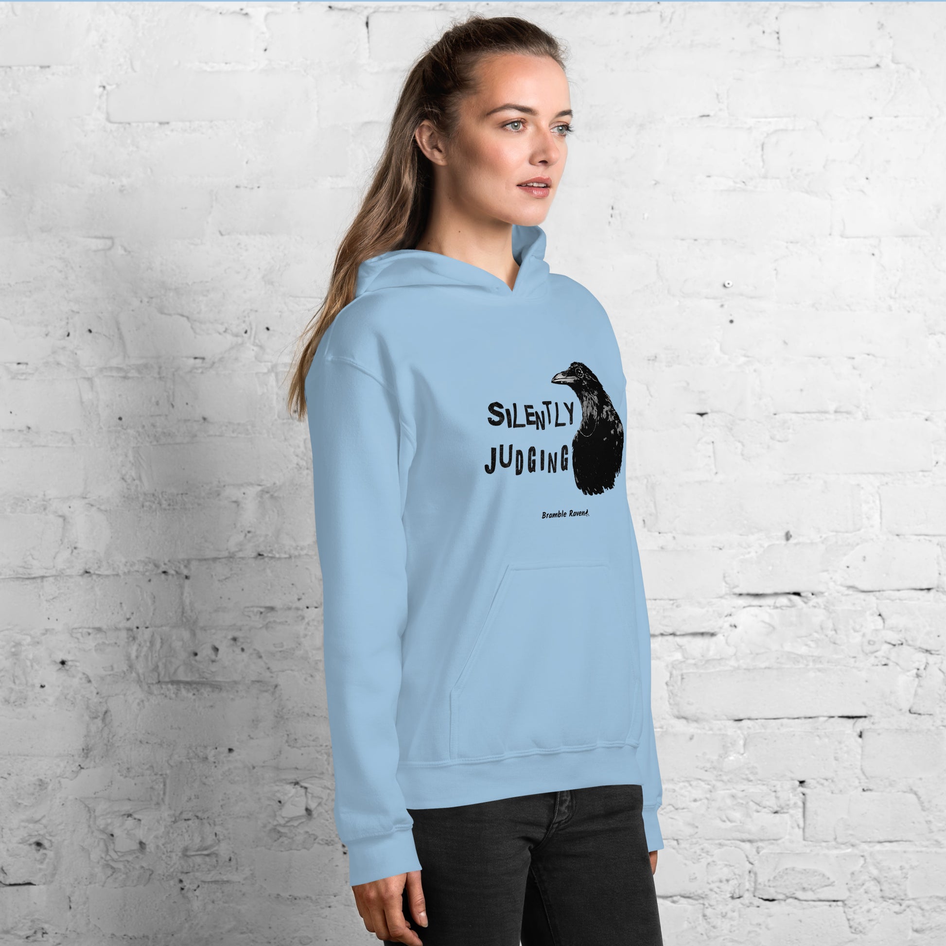 Unisex light blue colored hoodie with horizontal design of silently judging text by black crow wearing a monocle.  Design on the front of hoodie. Features double-lined hood and front pouch pocket. Shown on female model facing right.
