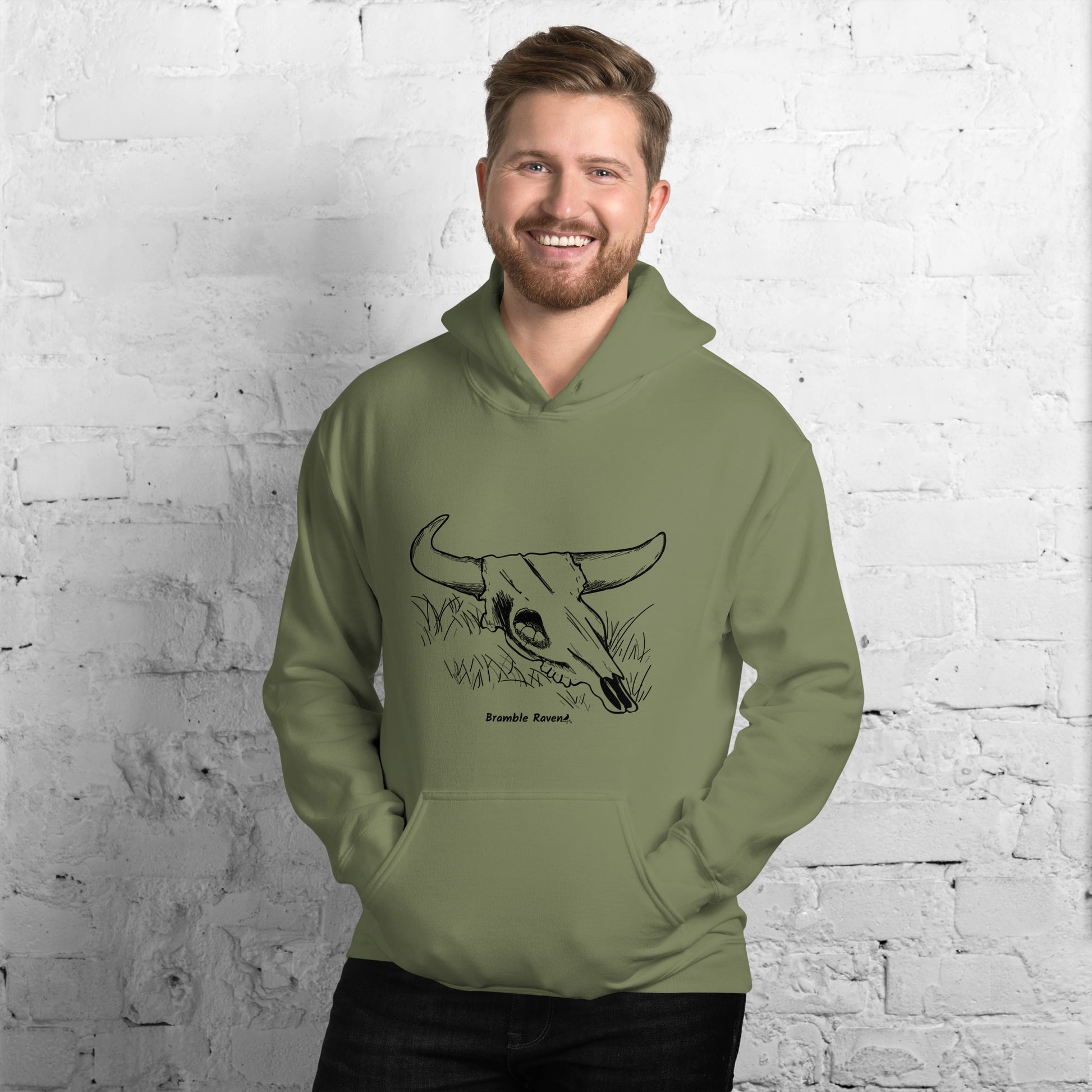 Military green colored unisex hoodie. Front has image of a cow skull cradling a bird nest. Features double-lined hood and front pouch pocket. Shown on male model in front of brick wall.