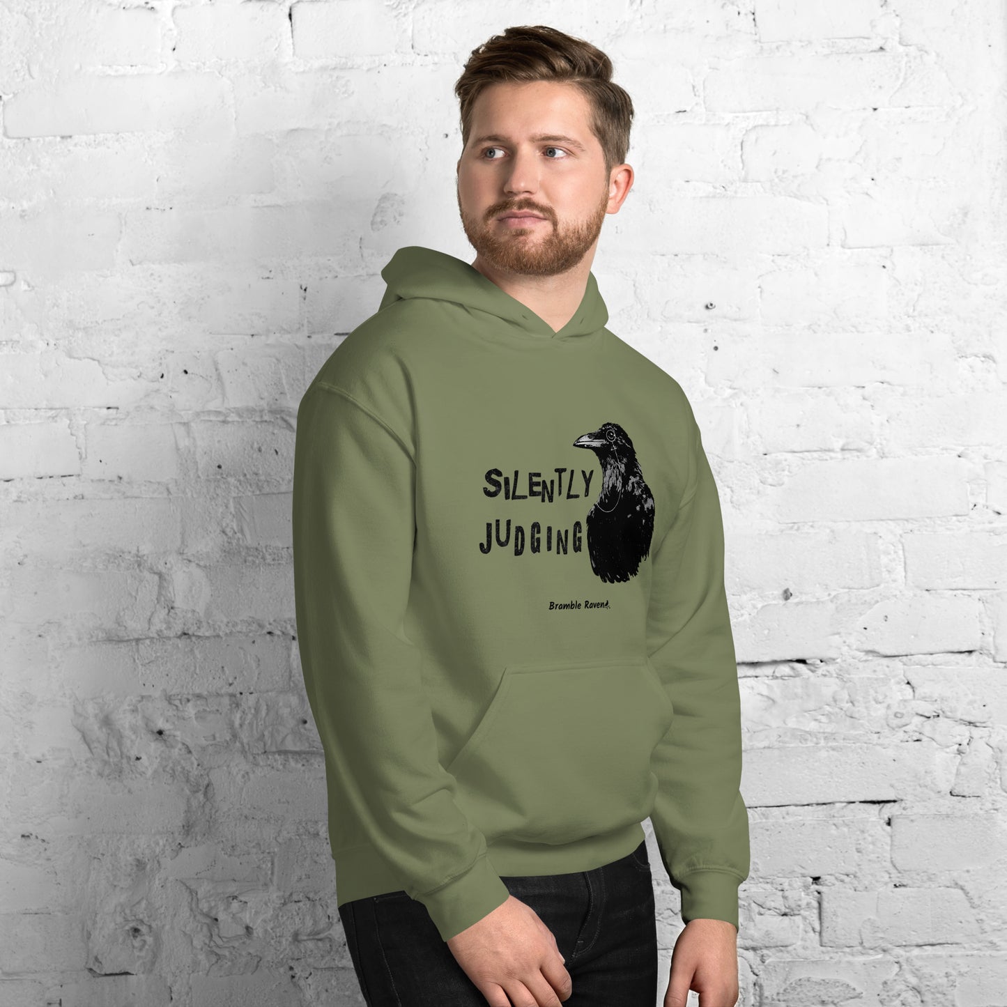 Unisex military green colored hoodie with horizontal design of silently judging text by black crow wearing a monocle.  Design on the front of hoodie. Features double-lined hood and front pouch pocket. Shown on male model.