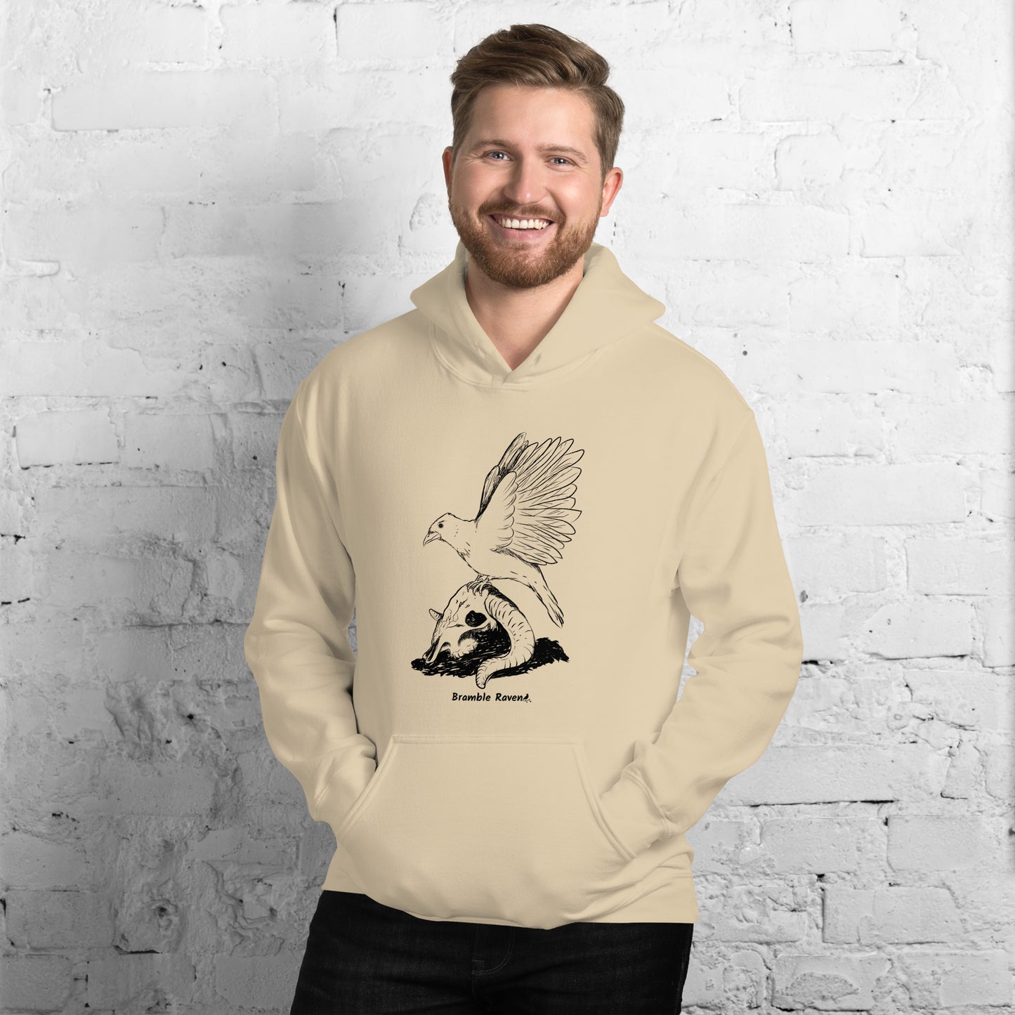 Sand colored unisex Reflections hoodie. Features image of a crow with wings outstretched sitting on a sheep skull.  Has a front pouch pocket and double lined hood. Shown on male model by white brick wall.
