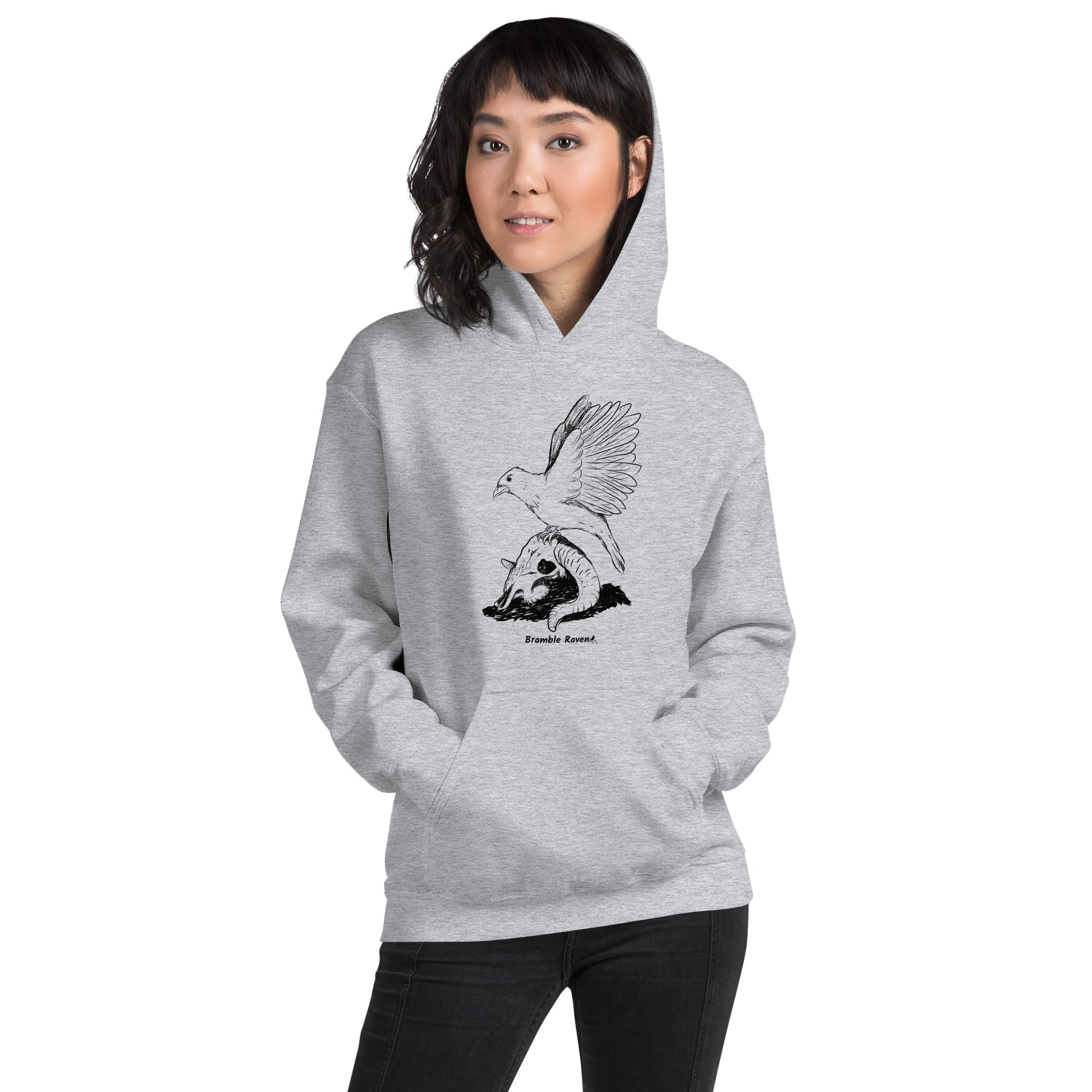 Sport grey colored unisex Reflections hoodie. Features image of a crow with wings outstretched sitting on a sheep skull.  Has a front pouch pocket and double lined hood. Shown on female model.
