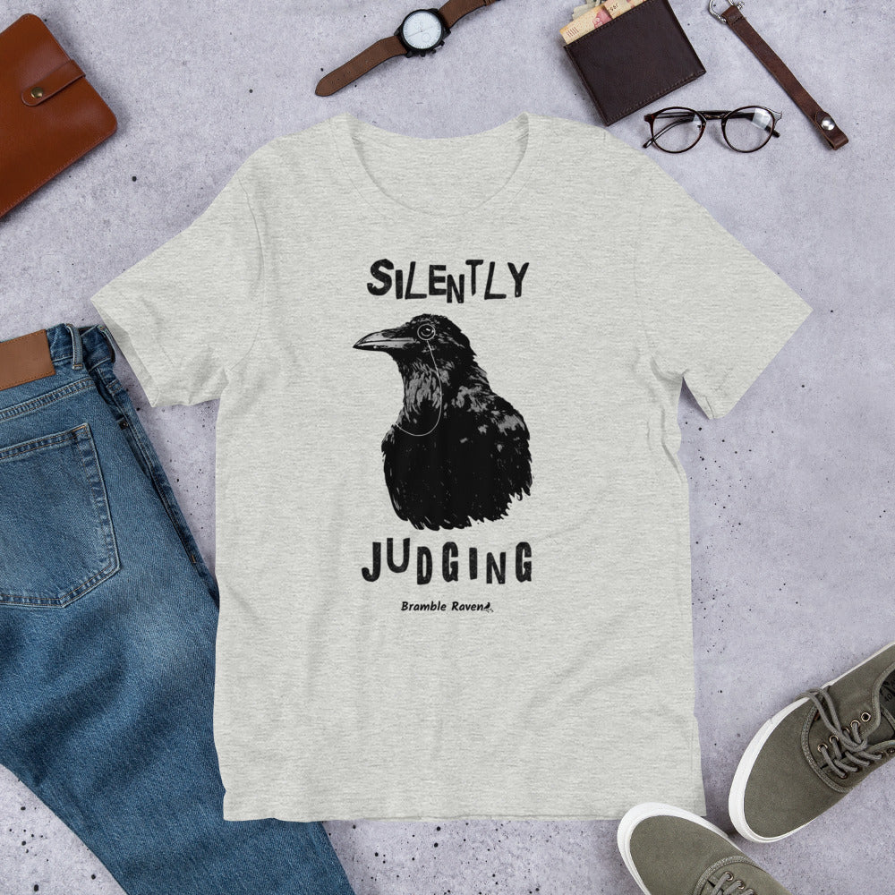 Unisex athletic heather grey colored t-shirt. Features vertical image of silently judging text above and below black crow wearing a monocle.  Shown on ground by pants, shoes, glasses, wallet, and watch.