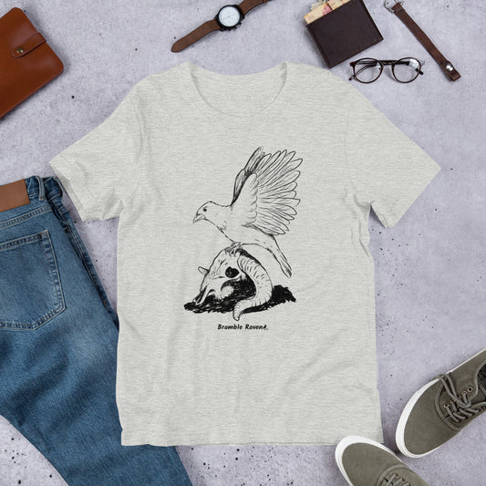 Athletic heather grey colored unisex t-shirt. Features Reflections illustration of a crow with wings outstretched sitting on a sheep skull. Shown on floor by pants and shoes.