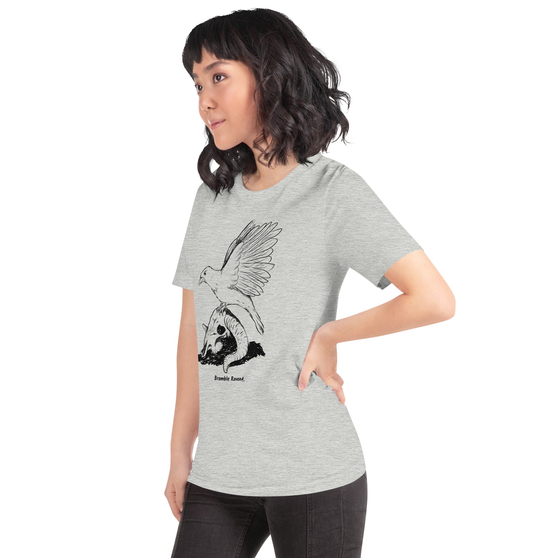 Athletic heather grey colored unisex t-shirt. Features Reflections illustration of a crow with wings outstretched sitting on a sheep skull. Shown on female model.