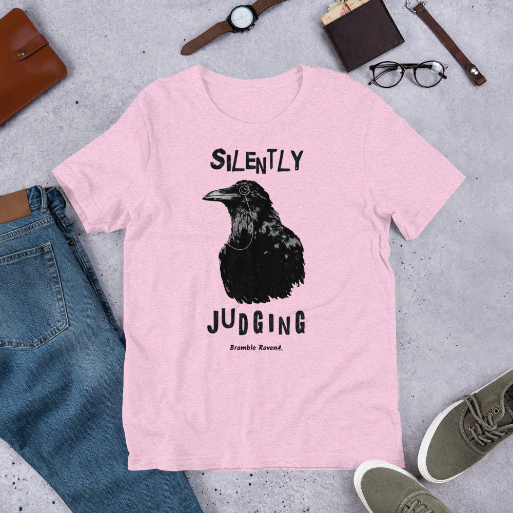 Unisex heather prism lilac pink colored t-shirt. Features vertical image of silently judging text above and below black crow wearing a monocle.  Shown on ground by pants, shoes, glasses, wallet, and watch.