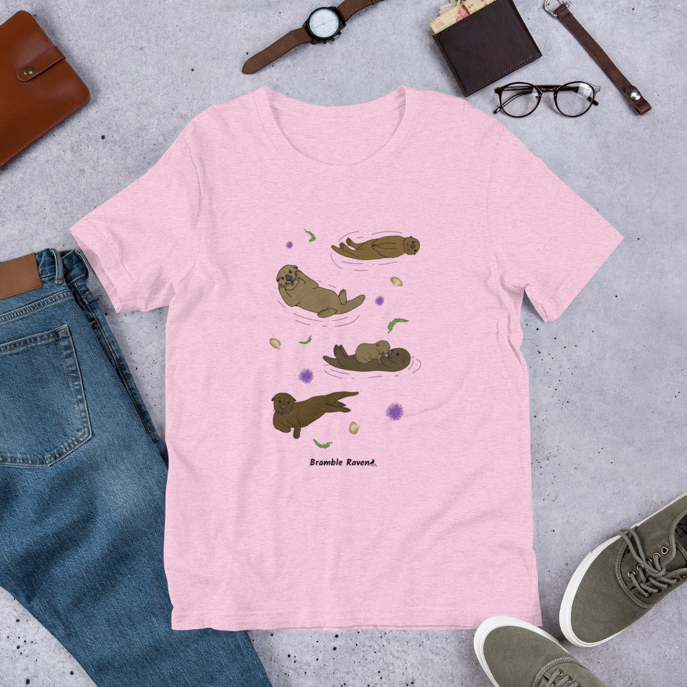 Unisex heather prism lilac pink colored t-shirt. Features four sea otters with seaweed, shells, and sea urchins. Shown surrounded by pants, shoes, and a wallet.