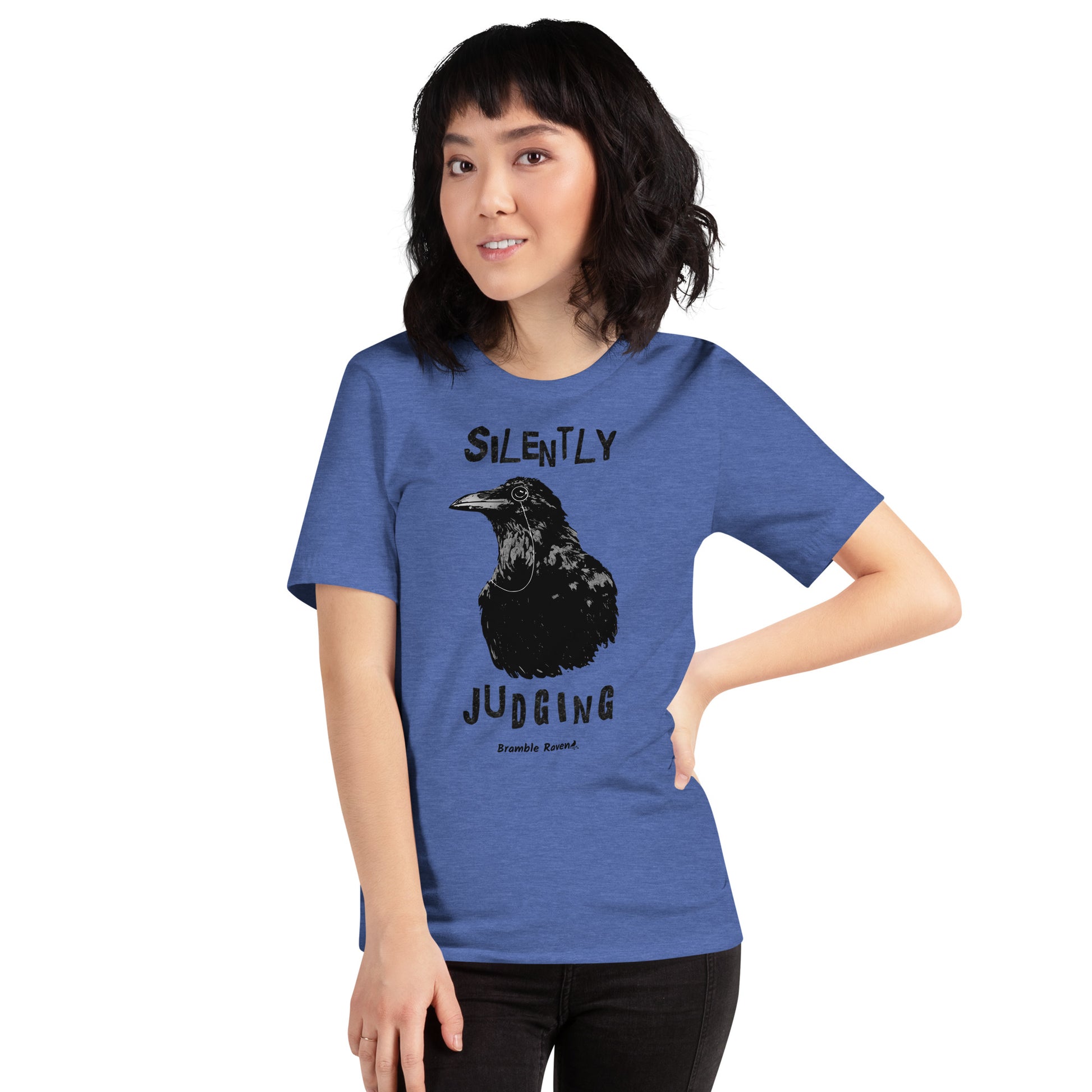 Unisex heather true royal blue colored t-shirt. Features vertical image of silently judging text above and below black crow wearing a monocle.  Shown on female model.