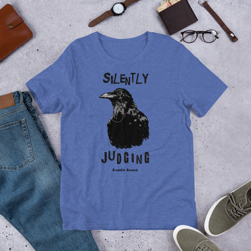 Unisex heather true royal blue colored t-shirt. Features vertical image of silently judging text above and below black crow wearing a monocle.  Shown on ground by pants, shoes, glasses, wallet, and watch.