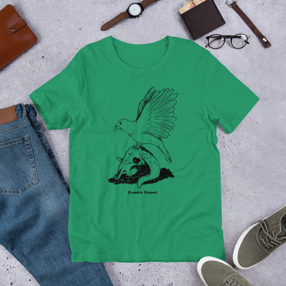 Kelly green colored unisex t-shirt. Features Reflections illustration of a crow with wings outstretched sitting on a sheep skull. Shown on floor by pants and shoes.