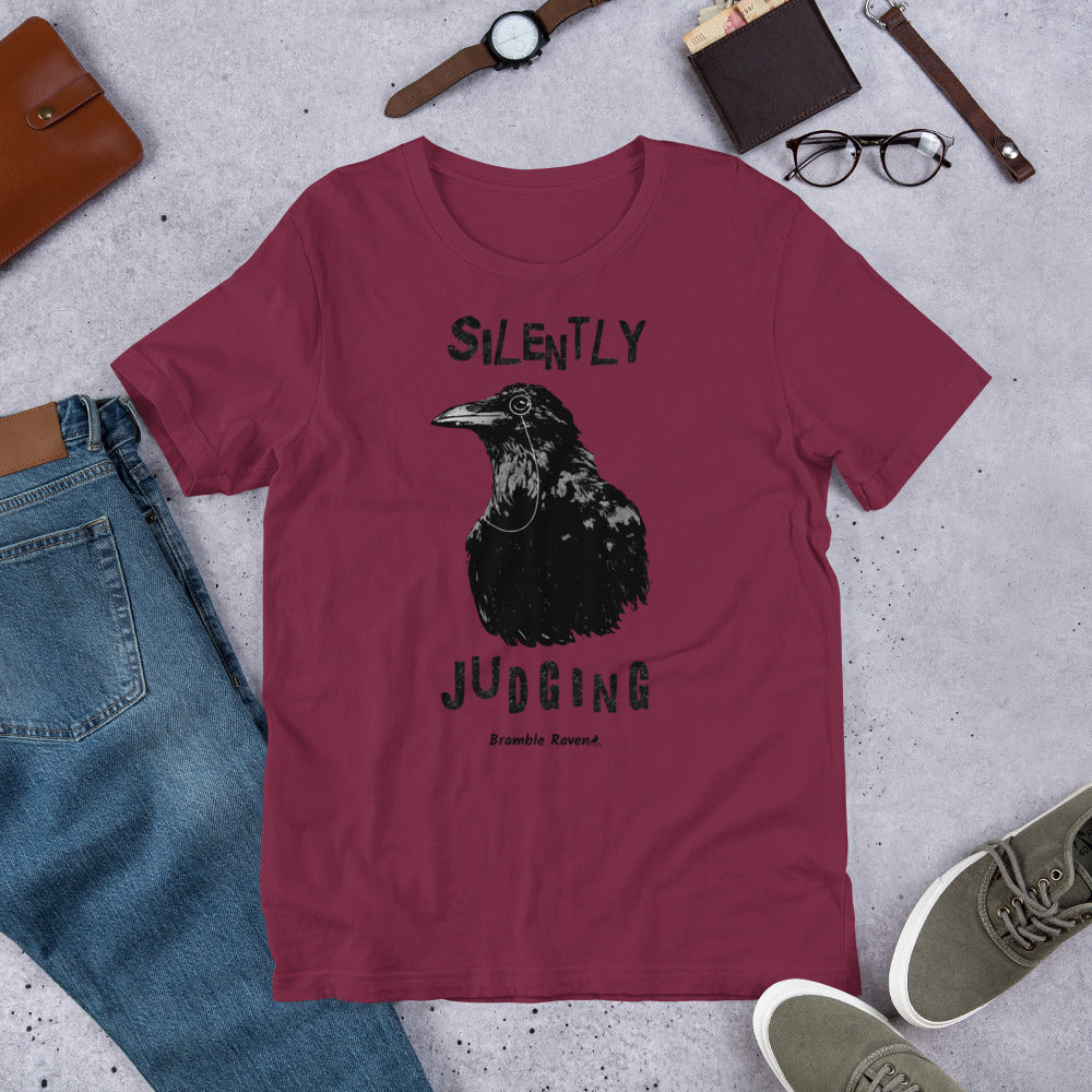 Unisex maroon colored t-shirt. Features vertical image of silently judging text above and below black crow wearing a monocle.  Shown on ground by pants, shoes, glasses, wallet, and watch.