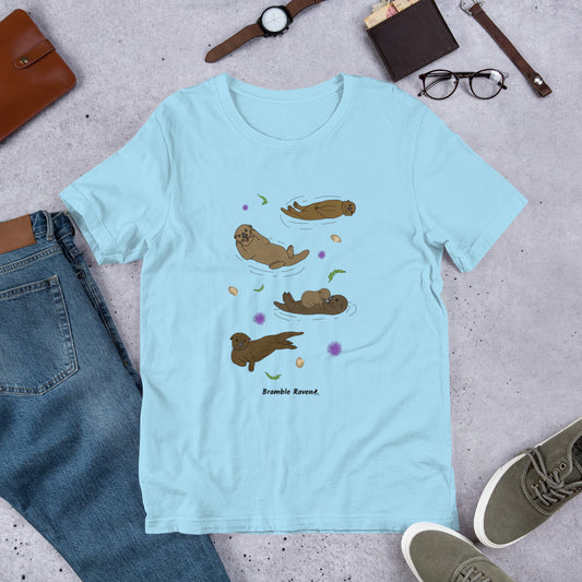 Unisex ocean blue colored t-shirt. Features four sea otters with seaweed, shells, and sea urchins. Shown surrounded by pants, shoes, and a wallet.