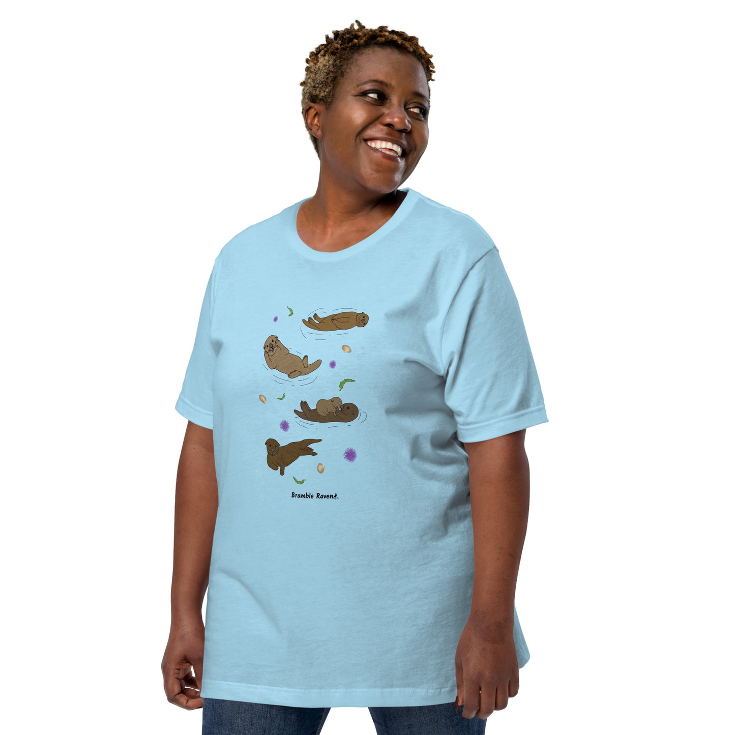 Unisex ocean blue colored t-shirt. Features four sea otters with seaweed, shells, and sea urchins. Shown on female model looking right.