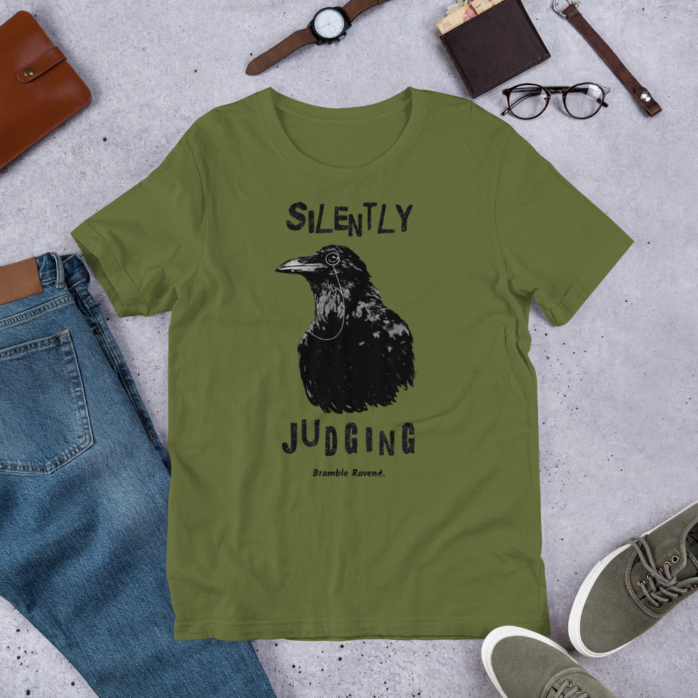Unisex olive green colored t-shirt. Features vertical image of silently judging text above and below black crow wearing a monocle.  Shown on ground by pants, shoes, glasses, wallet, and watch.