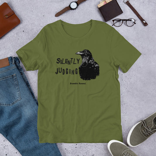 Unisex olive green colored t-shirt. Features horizontal image of silently judging text next to black crow wearing a monocle.  Shown on ground by pants, shoes, glasses, wallet, and watch.