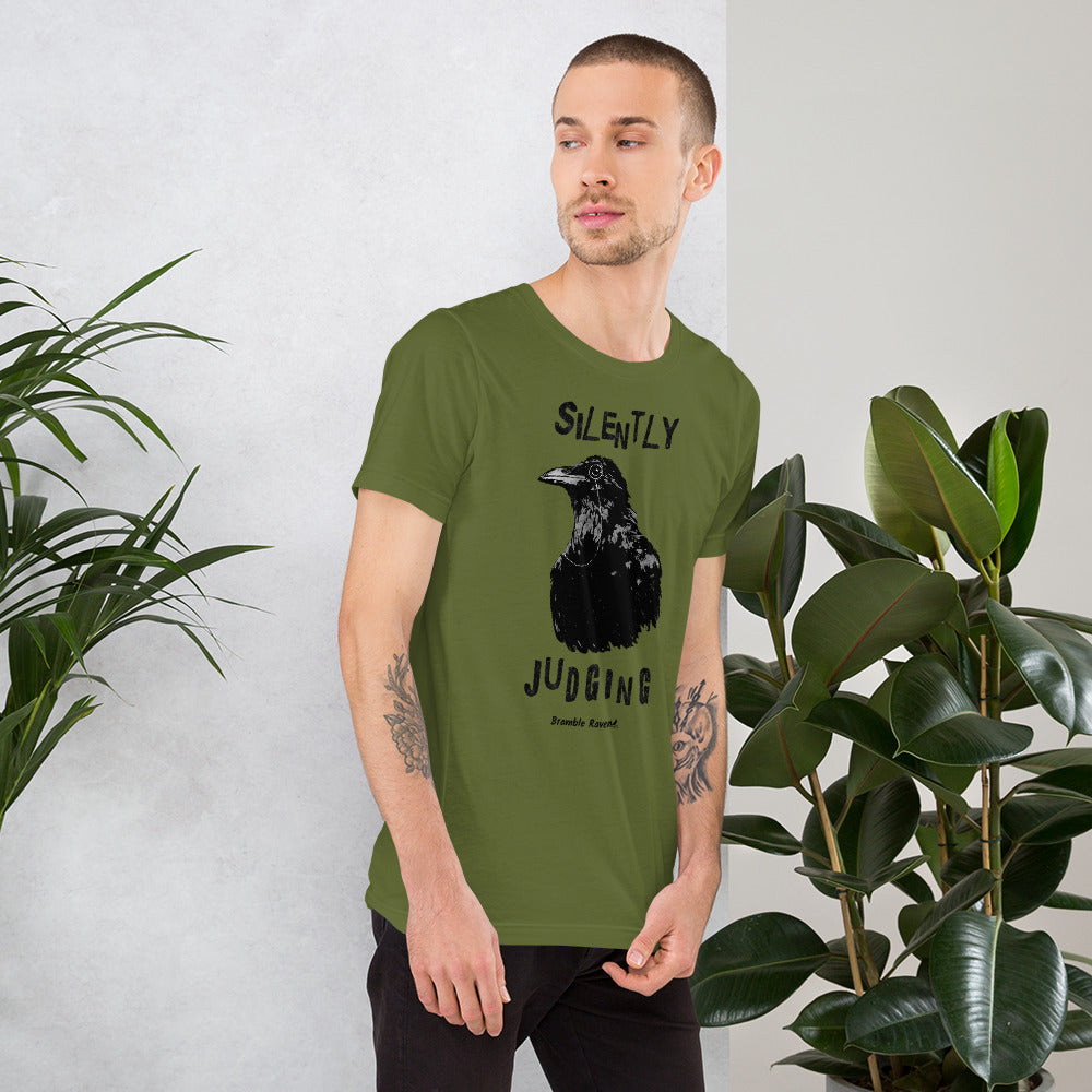 Unisex olive green colored t-shirt. Features vertical image of silently judging text above and below black crow wearing a monocle.  Shown on male model by houseplants.