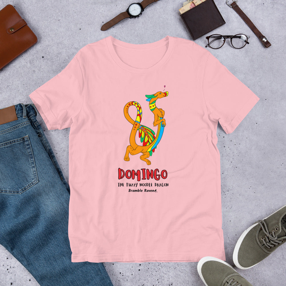 Domingo the Fuzzy Noodle Dragon on a pink unisex t-shirt. Shown surrounded by pants, shoes, glasses, a watch, and wallet.