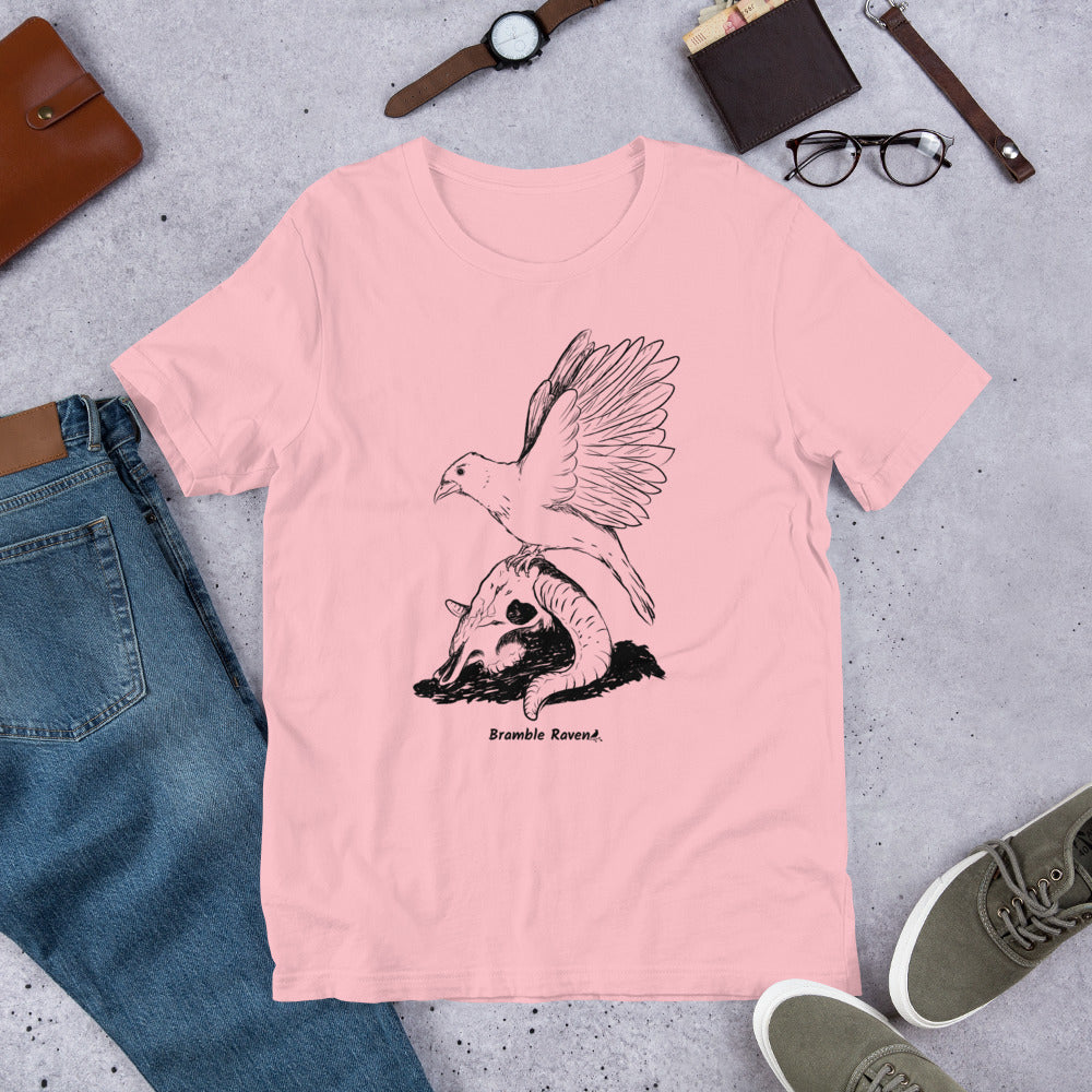 Pink colored unisex t-shirt. Features Reflections illustration of a crow with wings outstretched sitting on a sheep skull. Shown on floor by pants and shoes.