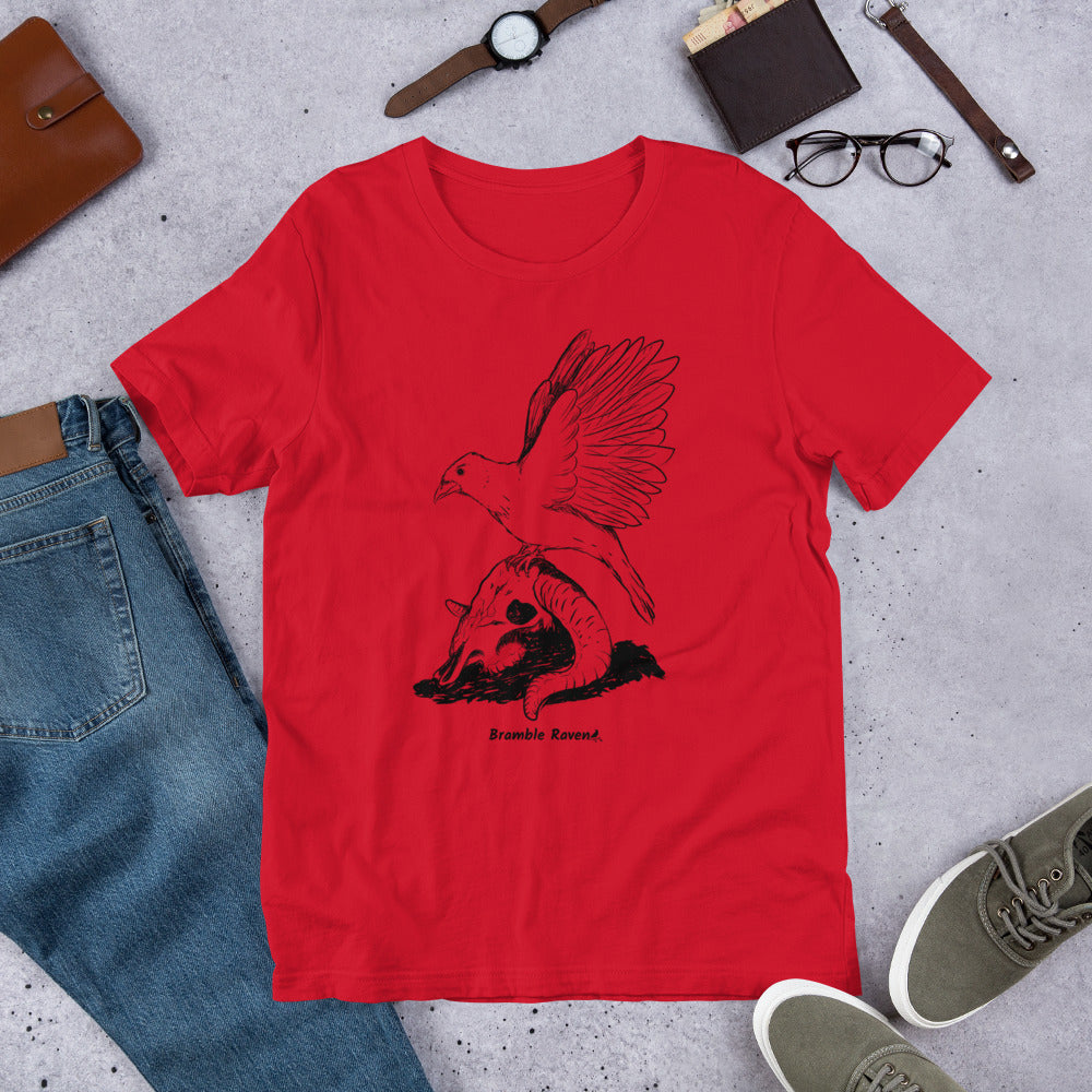 Red colored unisex t-shirt. Features Reflections illustration of a crow with wings outstretched sitting on a sheep skull. Shown on floor by pants and shoes.