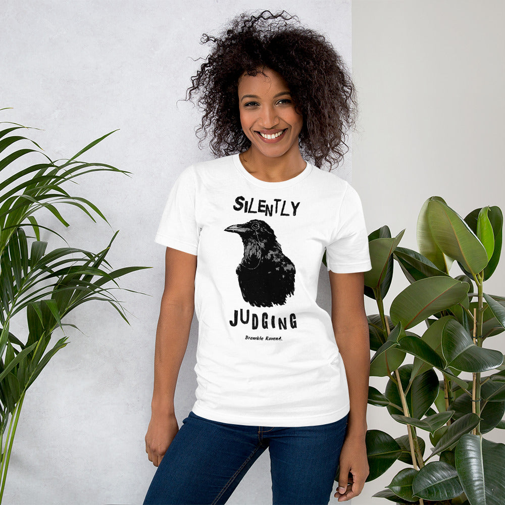 Unisex white colored t-shirt. Features vertical image of silently judging text above and below black crow wearing a monocle.  Shown on female model by houseplants.