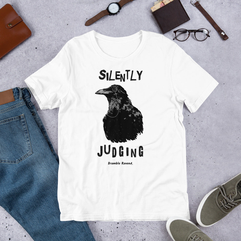 Unisex white colored t-shirt. Features vertical image of silently judging text above and below black crow wearing a monocle.  Shown on ground by pants, shoes, glasses, wallet, and watch.