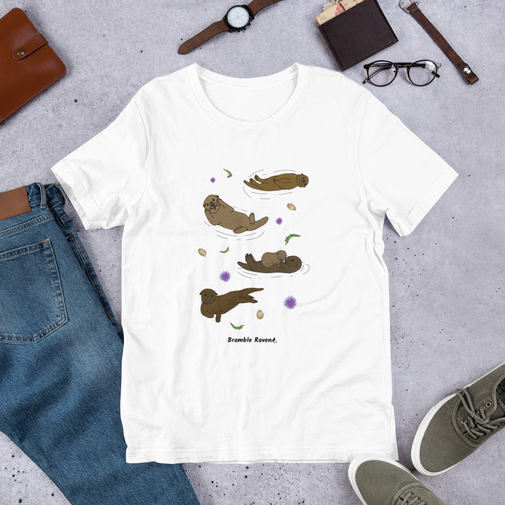 Unisex white colored t-shirt. Features four sea otters with seaweed, shells, and sea urchins. Shown surrounded by pants, shoes, and a wallet.