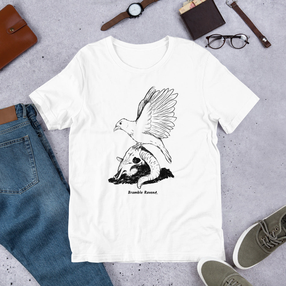 White colored unisex t-shirt. Features Reflections illustration of a crow with wings outstretched sitting on a sheep skull. Shown on floor by pants and shoes.