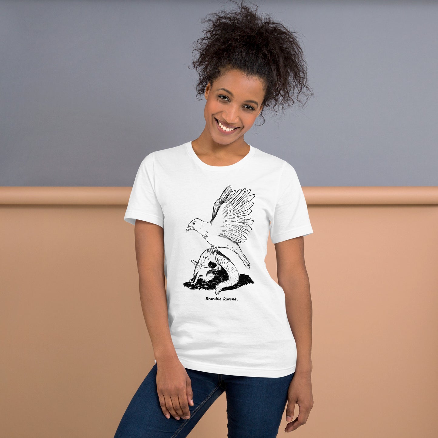 White colored unisex t-shirt. Features Reflections illustration of a crow with wings outstretched sitting on a sheep skull. Shown on female model against tan wall.