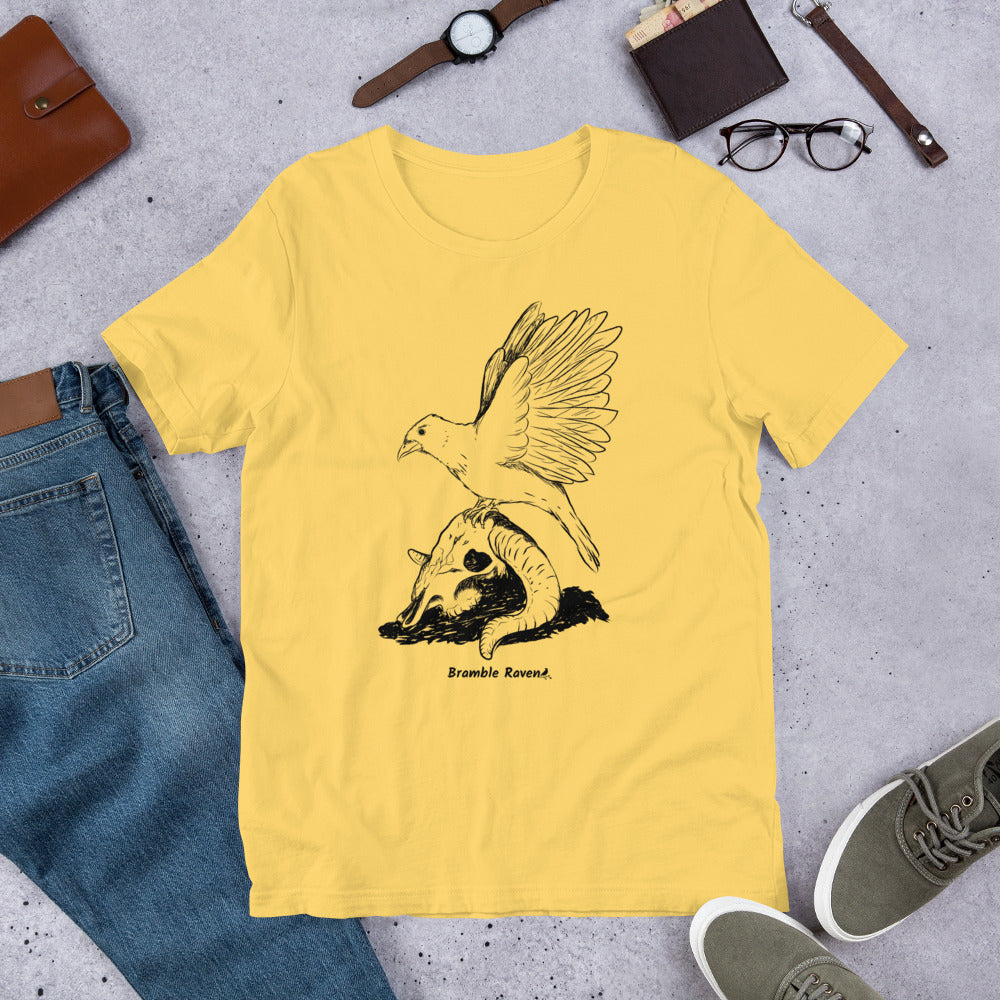 Yellow colored unisex t-shirt. Features Reflections illustration of a crow with wings outstretched sitting on a sheep skull. Shown on floor by pants and shoes.