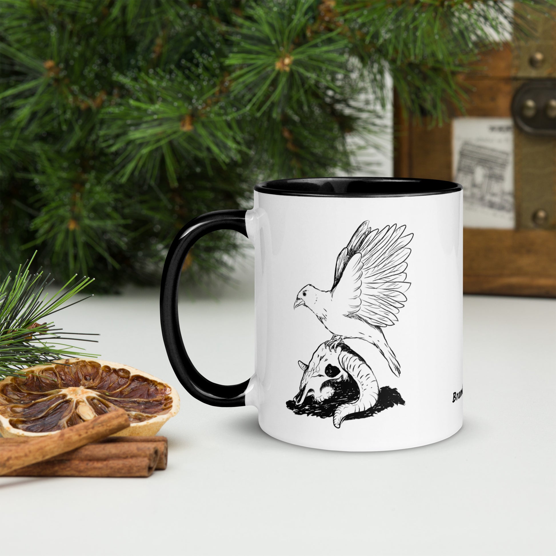 11 ounce white ceramic mug with black handle and inside. Features a double sided print of Reflections, a design with a crow sitting on a sheep skull. Dishwasher and microwave safe. Shown by dried citrus and pine boughs.