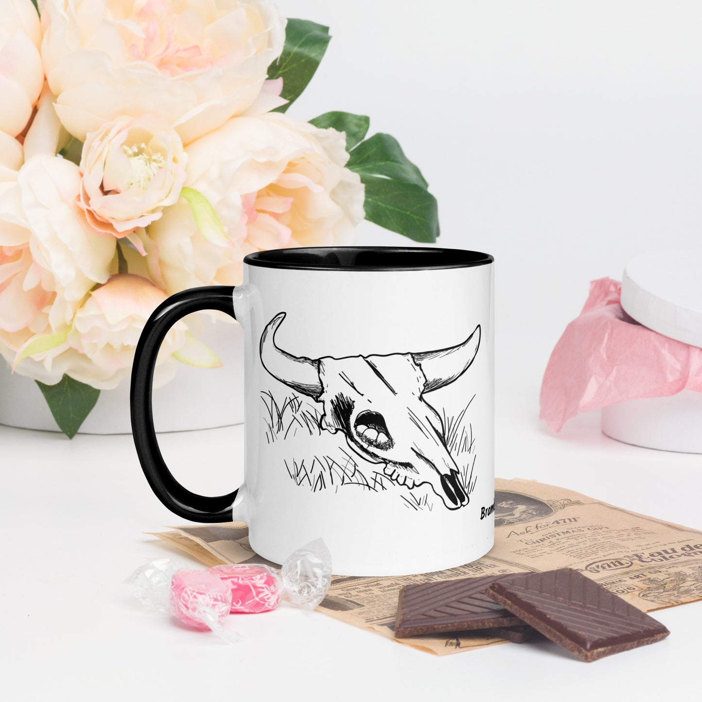 11 ounce ceramic mug. White with black handle and inside. Features double sided image of a cow skull cradling a bird nest. Shown by flowers and chocolates.