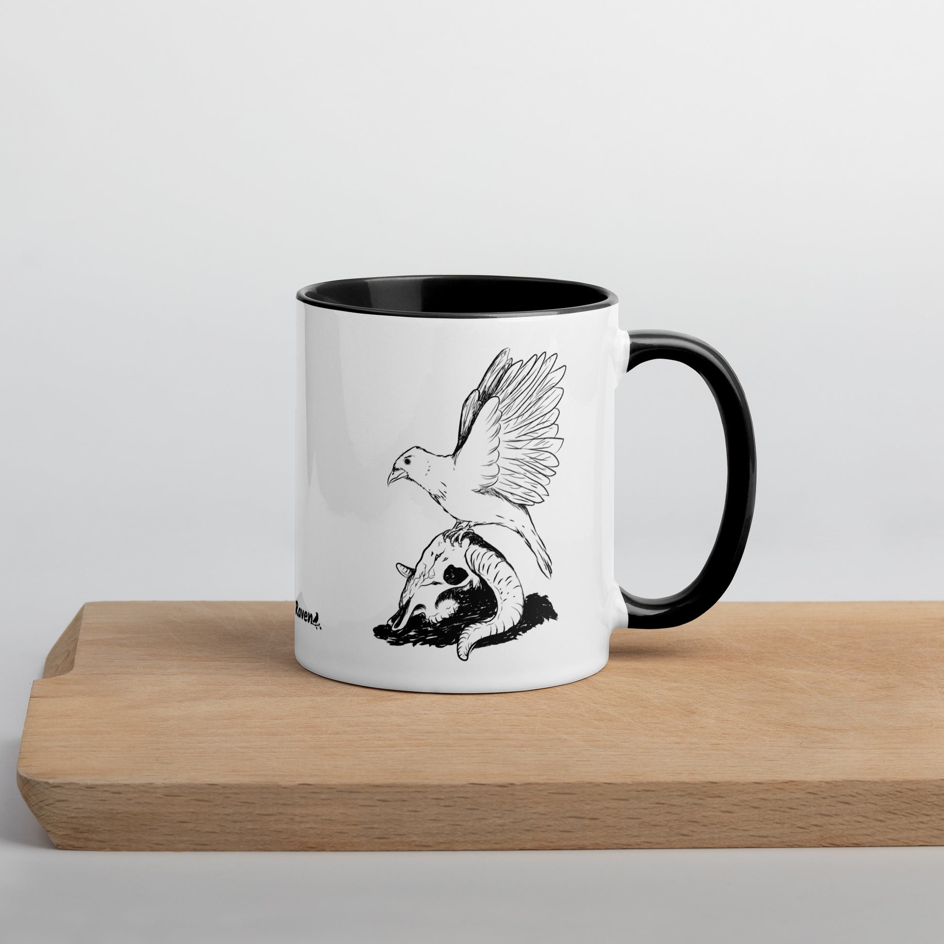 11 ounce white ceramic mug with black handle and inside. Features a double sided print of Reflections, a design with a crow sitting on a sheep skull. Dishwasher and microwave safe. Shown on wooden cutting board.