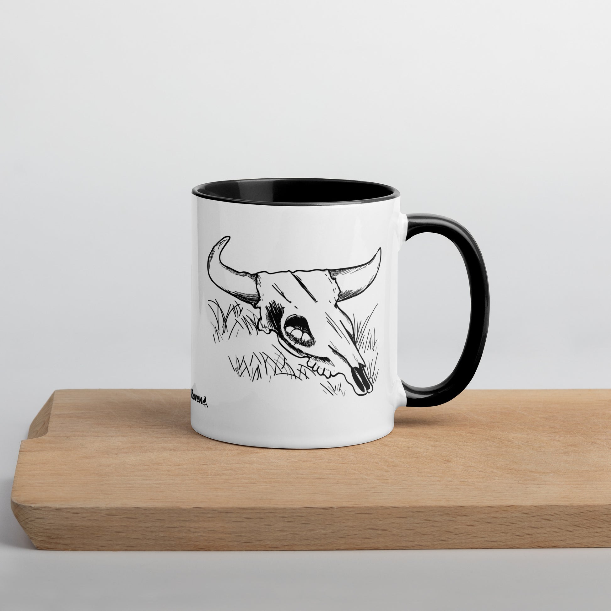11 ounce ceramic mug. White with black handle and inside. Features double sided image of a cow skull cradling a bird nest. Shown on wooden cutting board.