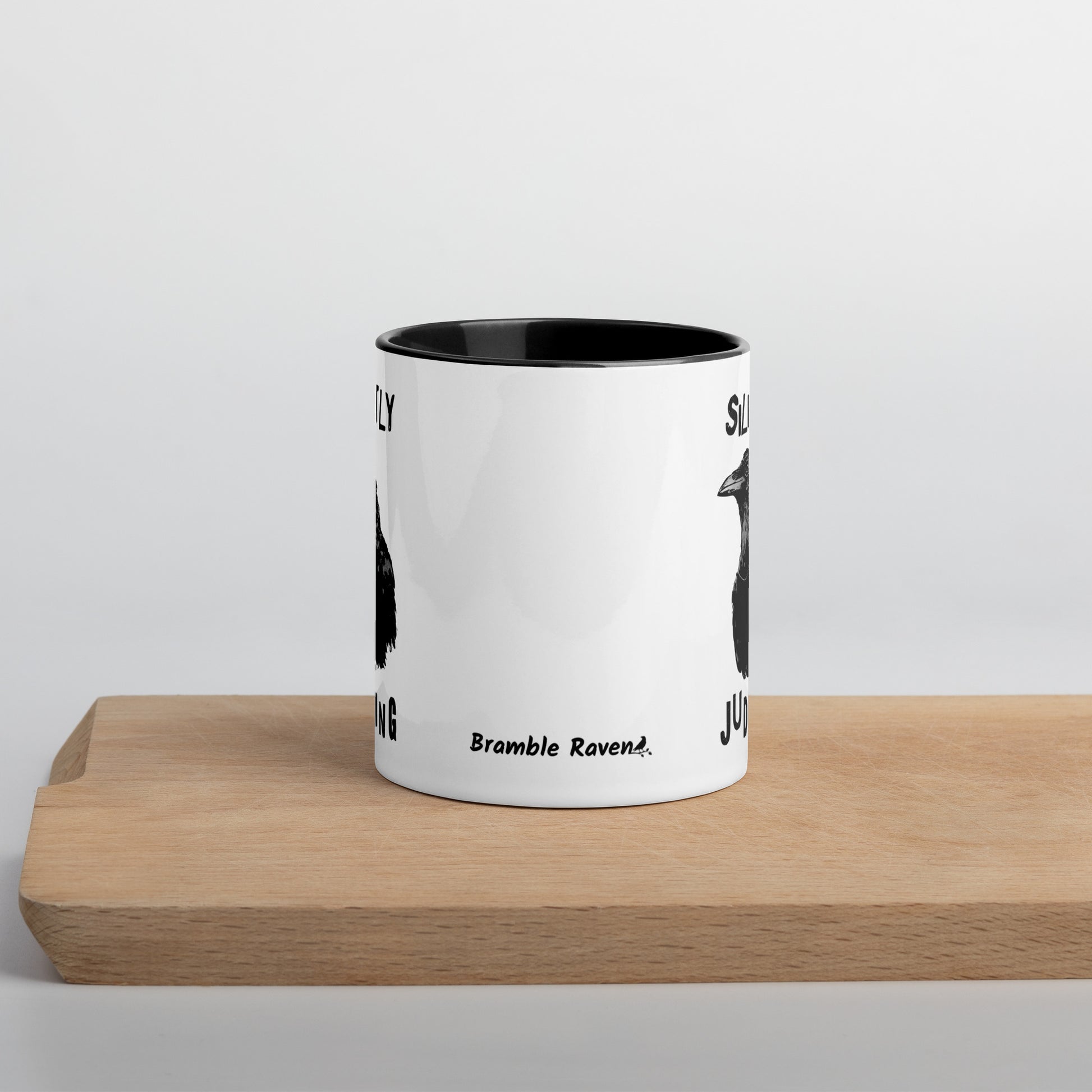 11 ounce ceramic mug with Silently Judging text surrounding a monacle-wearing crow. Double-sided design. Mug has black rim, handle, and inside. Front of mug shown sitting on wooden cutting board.