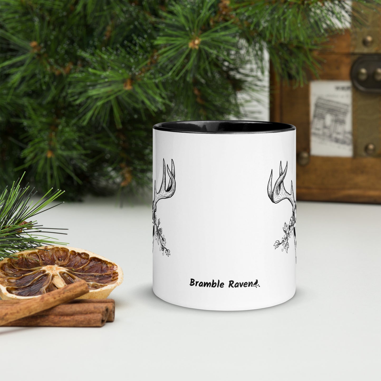11 ounce ceramic mug with hand illustrated deer skull wreathed in flowers. Double-sided design. Mug has black rim, handle, and inside. Front image of mug shows Bramble Raven logo. Shown sitting on a tabletop by pine branches, cinnamon sticks, and dried orange slice.