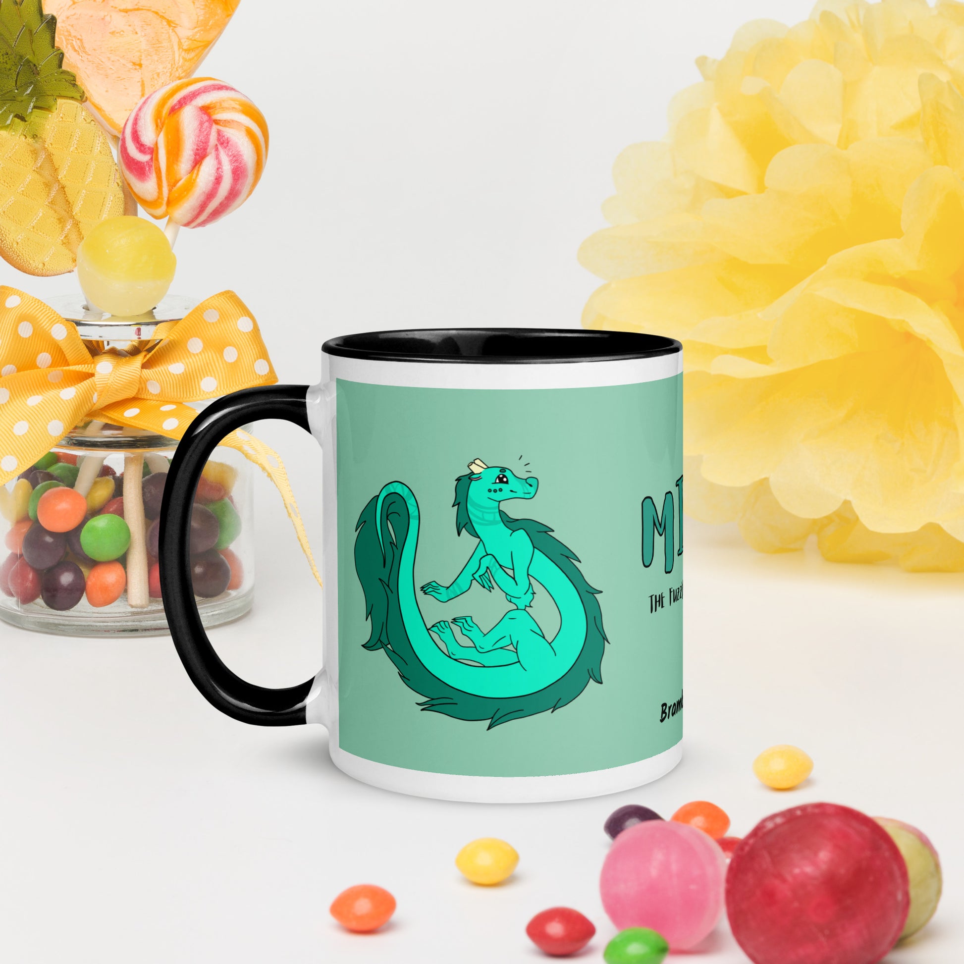 11 ounce white ceramic mug. Black inside and handle. Features double-sided image of Minty the Fuzzy Noodle Dragon on a green background. Shown on tabletop by candy and decorations.
