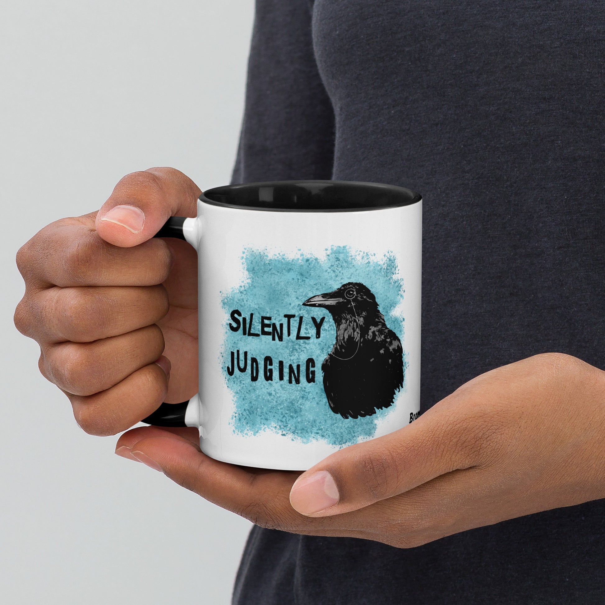 11 ounce ceramic mug with Silently Judging text next to a monacle-wearing crow on blue paint splatters. Double-sided design. Mug has black rim, handle, and inside. Shown in the hands of a model.