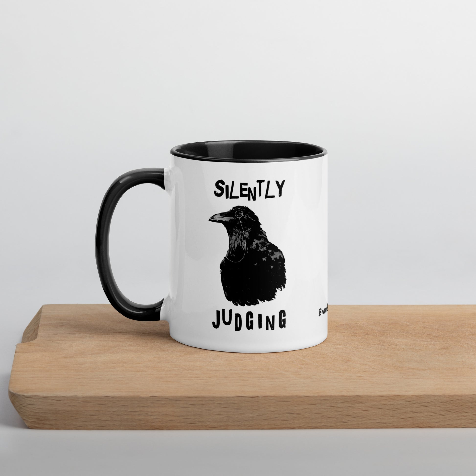 11 ounce ceramic mug with Silently Judging text surrounding a monacle-wearing crow. Double-sided design. Mug has black rim, handle, and inside. Shown sitting on wooden cutting board.