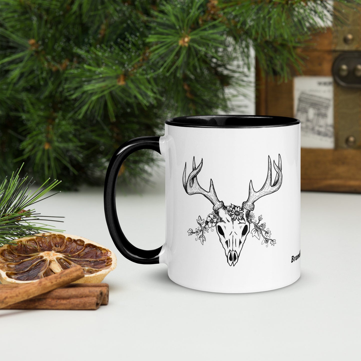 11 ounce ceramic mug with hand illustrated deer skull wreathed in flowers. Double-sided design. Mug has black rim, handle, and inside. Shown sitting on a tabletop by pine branches, cinnamon sticks, and dried orange slice.