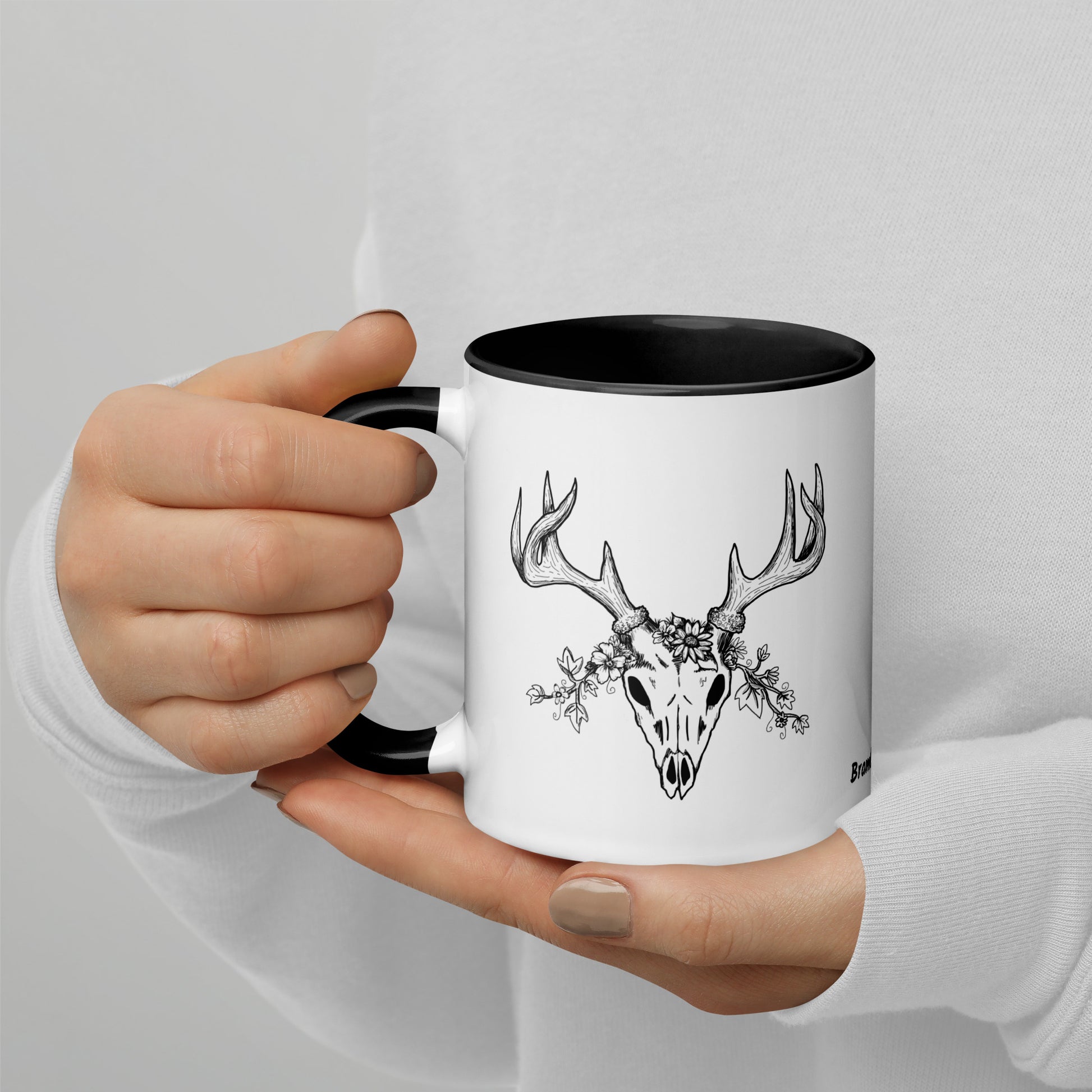 11 ounce ceramic mug with hand illustrated deer skull wreathed in flowers. Double-sided design. Mug has black rim, handle, and inside. Shown in model's hand.