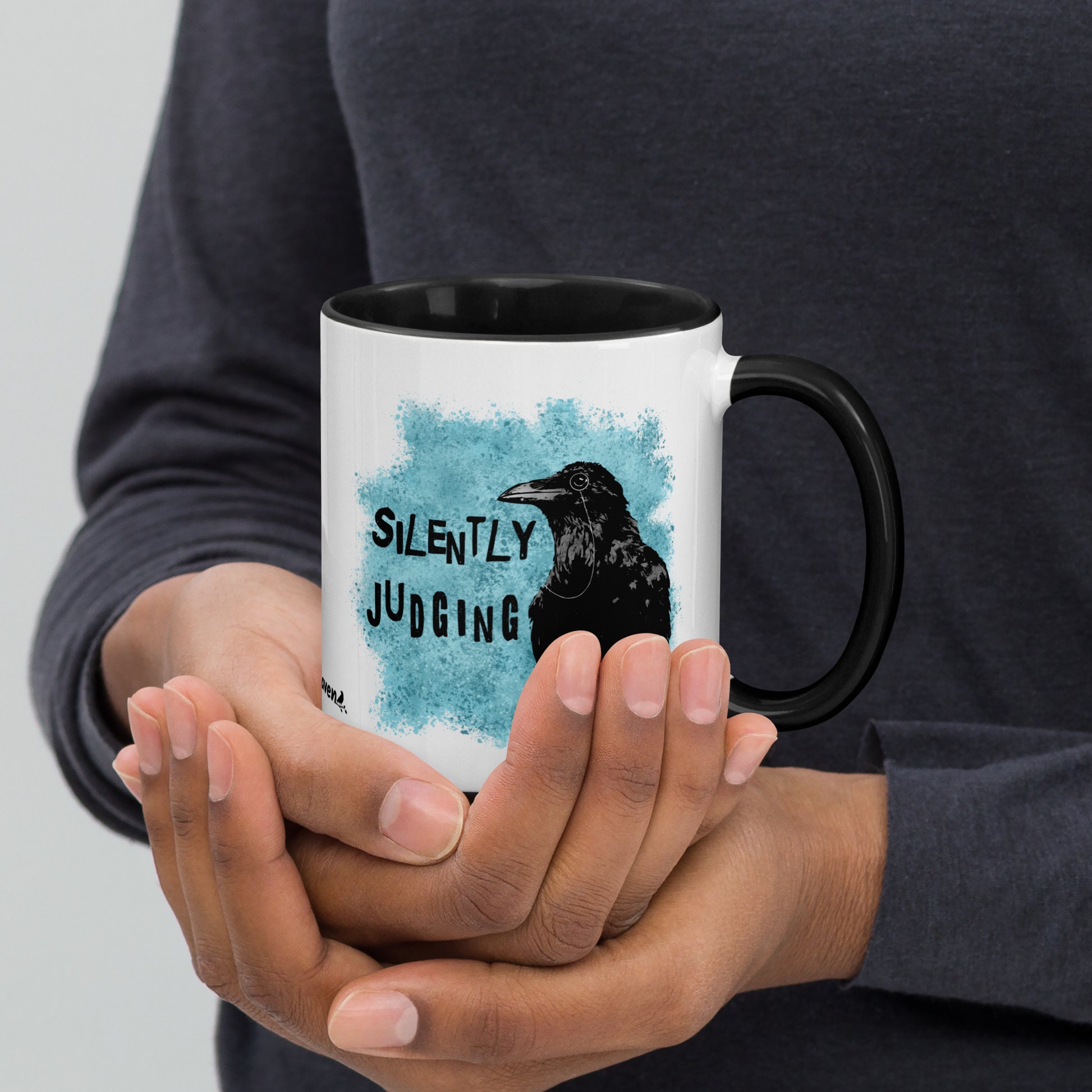 11 ounce ceramic mug with Silently Judging text next to a monacle-wearing crow on blue paint splatters. Double-sided design. Mug has black rim, handle, and inside. Shown in the hands of a model.