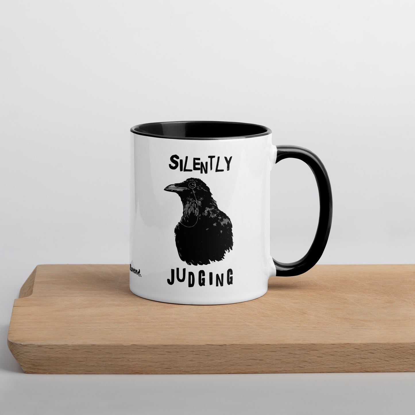 11 ounce ceramic mug with Silently Judging text surrounding a monacle-wearing crow. Double-sided design. Mug has black rim, handle, and inside. Shown sitting on wooden cutting board with handle facing right.