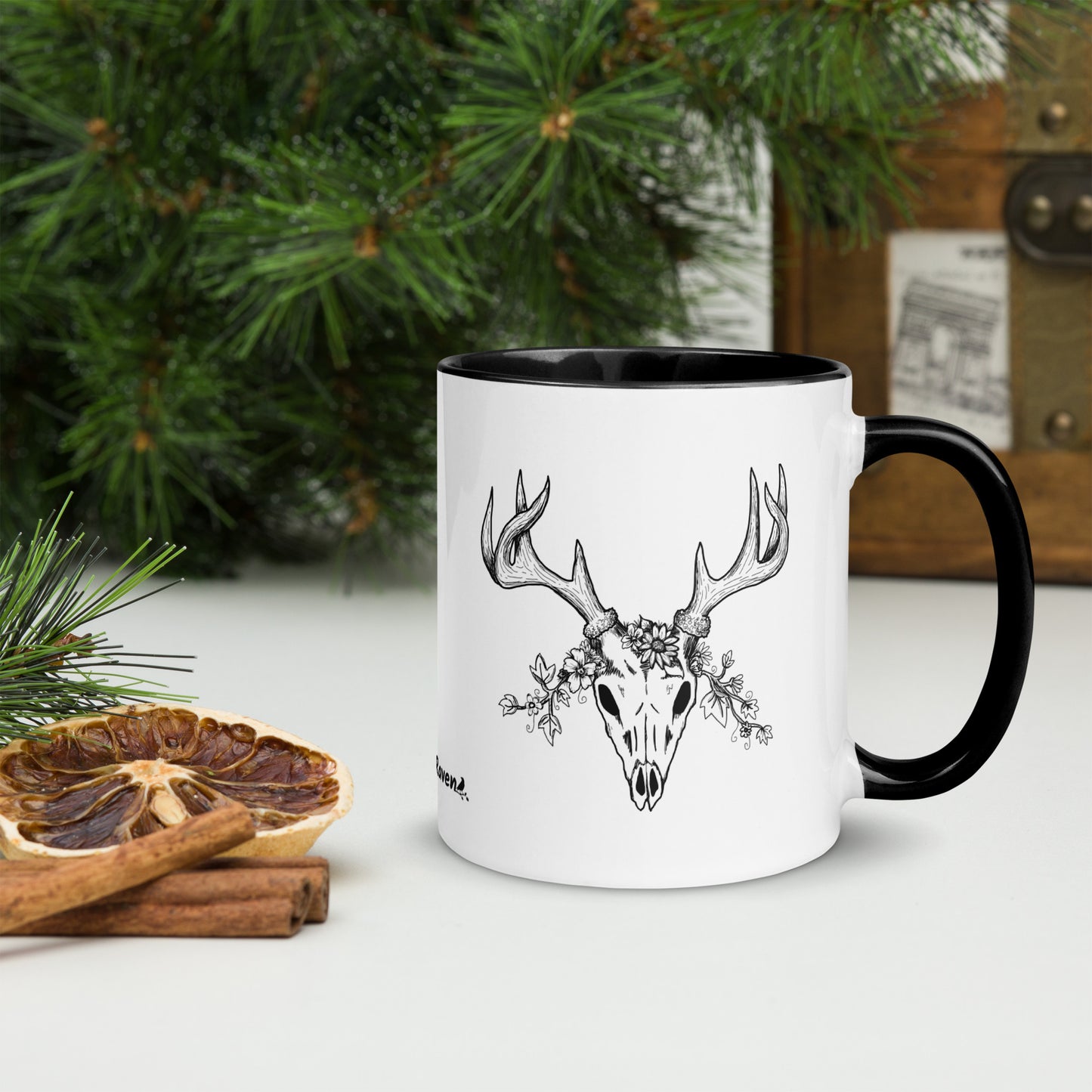 11 ounce ceramic mug with hand illustrated deer skull wreathed in flowers. Double-sided design. Mug has black rim, handle, and inside. Shown sitting on a tabletop by pine branches, cinnamon sticks, and dried orange slice.