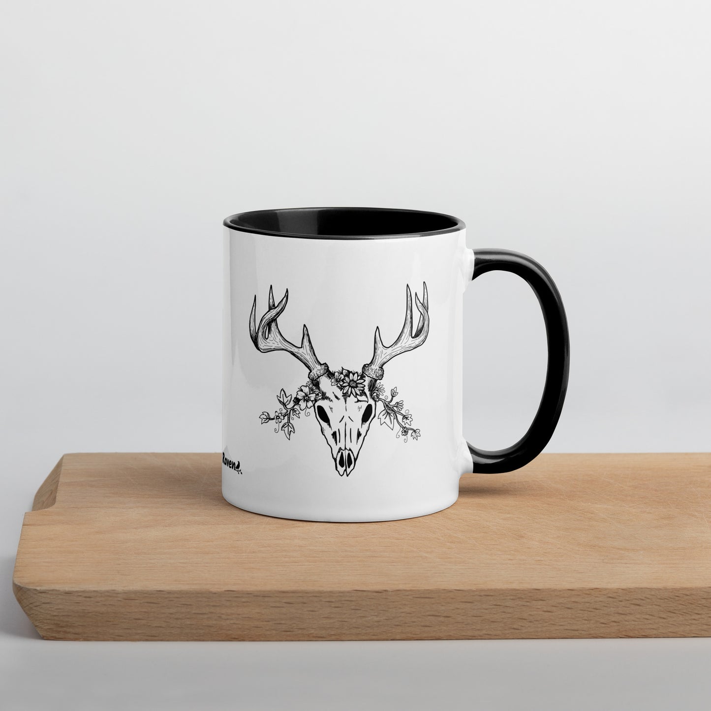 11 ounce ceramic mug with hand illustrated deer skull wreathed in flowers. Double-sided design. Mug has black rim, handle, and inside. Shown sitting on wooden cutting board with handle facing right.