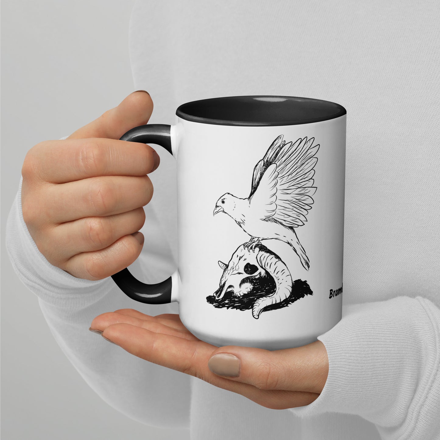 15 ounce white ceramic mug with black handle and inside. Features a double sided print of Reflections, a design with a crow sitting on a sheep skull. Dishwasher and microwave safe. Shown in model's hands.