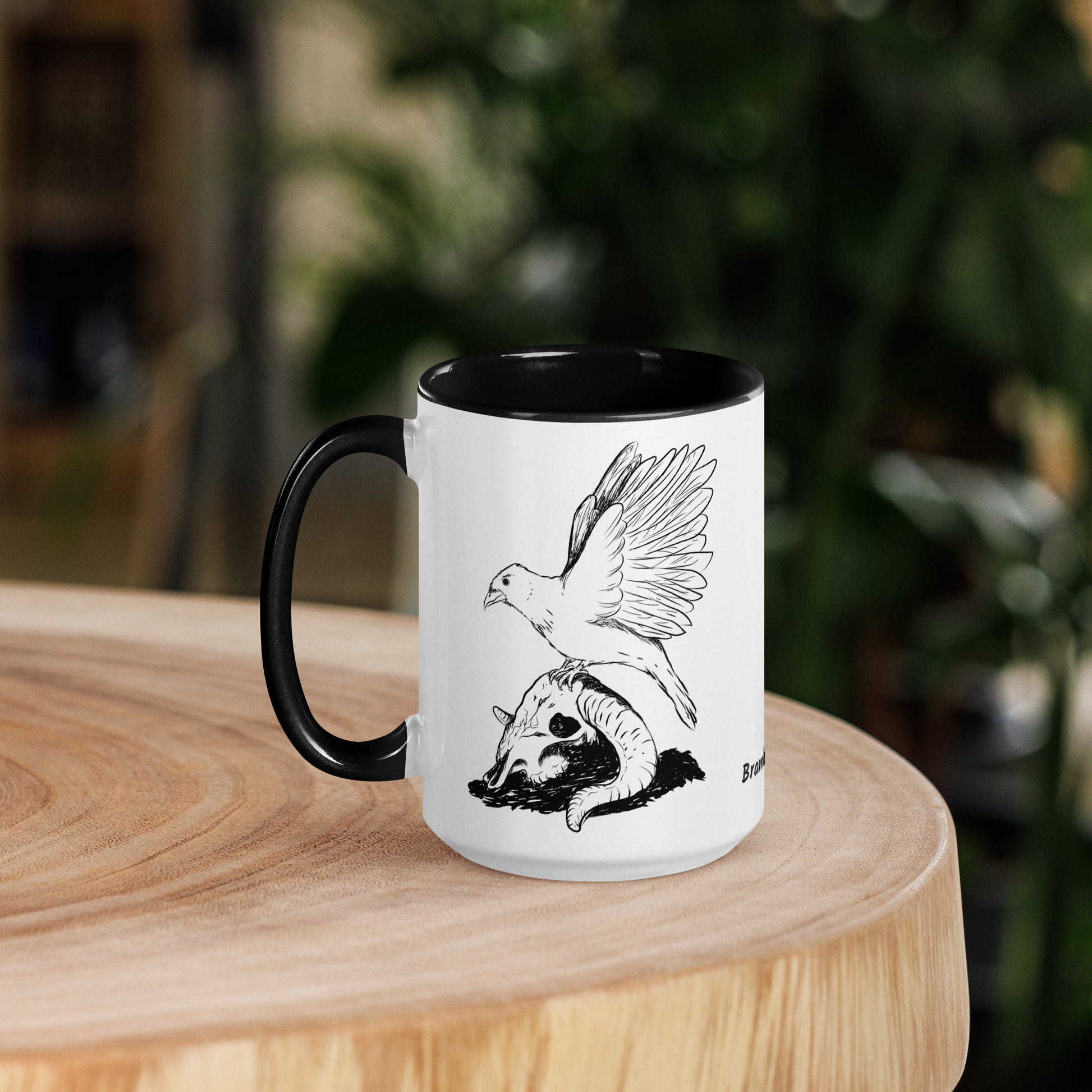 15 ounce white ceramic mug with black handle and inside. Features a double sided print of Reflections, a design with a crow sitting on a sheep skull. Dishwasher and microwave safe. Shown on wooden tabletop.