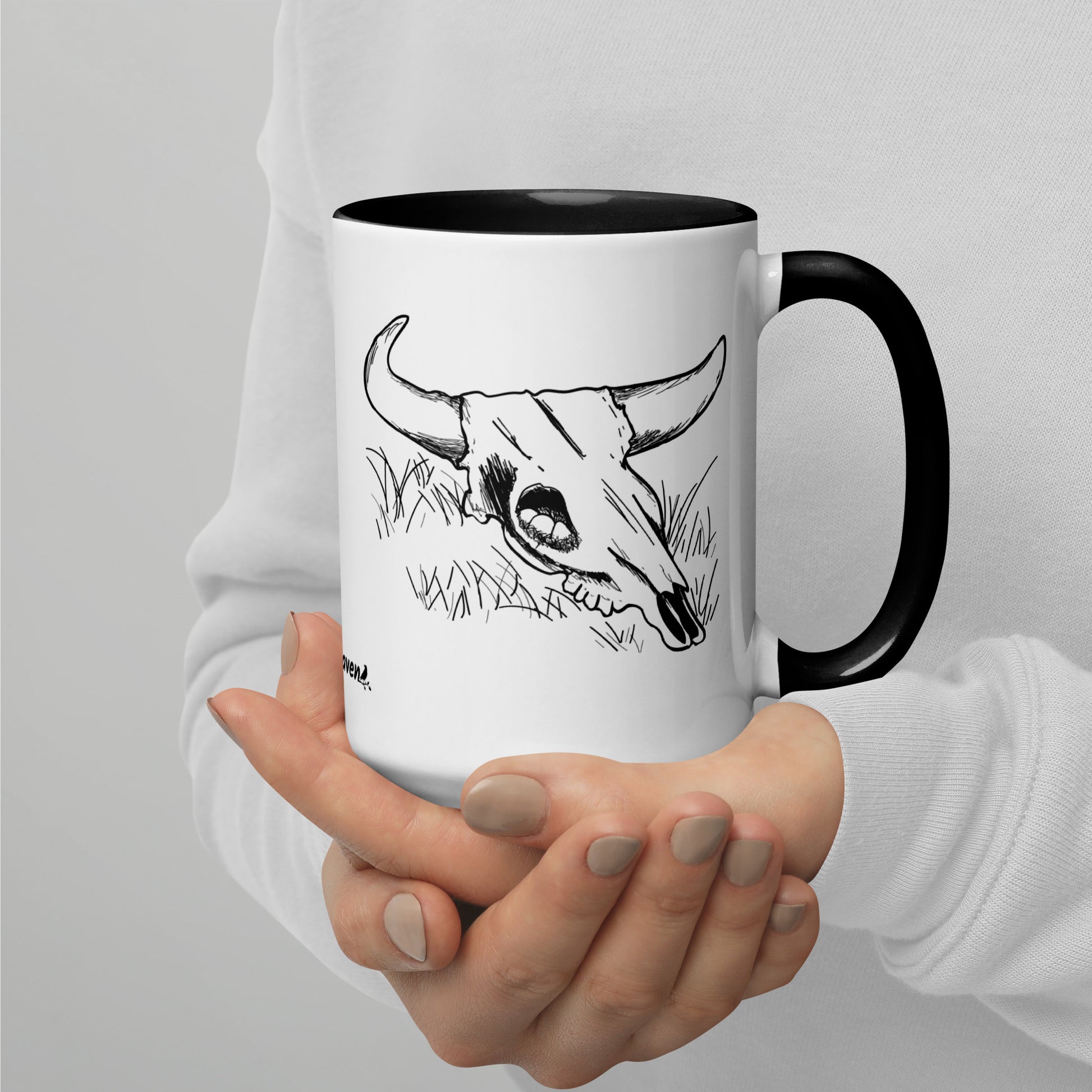 15 ounce ceramic mug. White with black handle and inside. Features double sided image of a cow skull cradling a bird nest. Shown in model's hand.