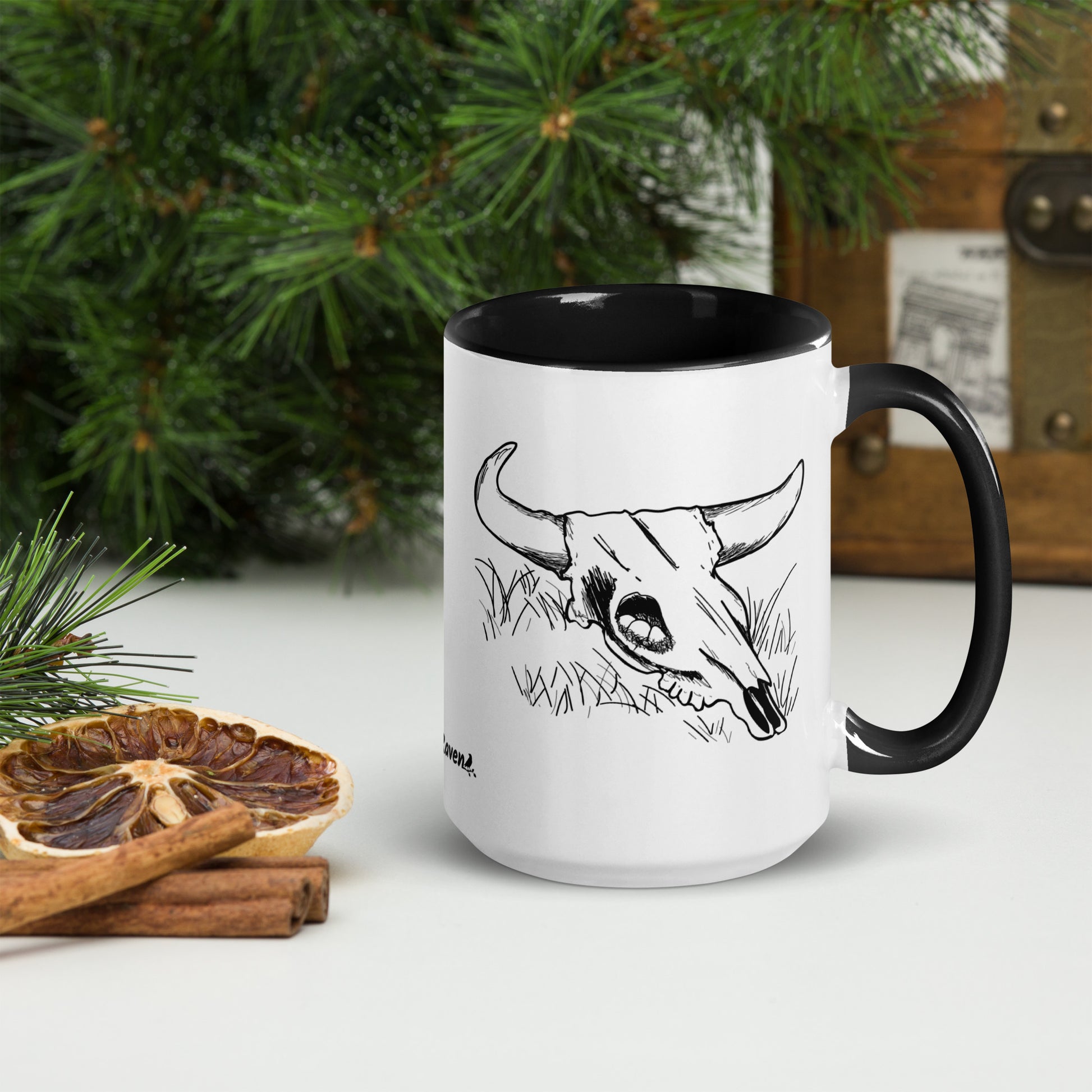 15 ounce ceramic mug. White with black handle and inside. Features double sided image of a cow skull cradling a bird nest. Shown by dried citrus slices and pine boughs.