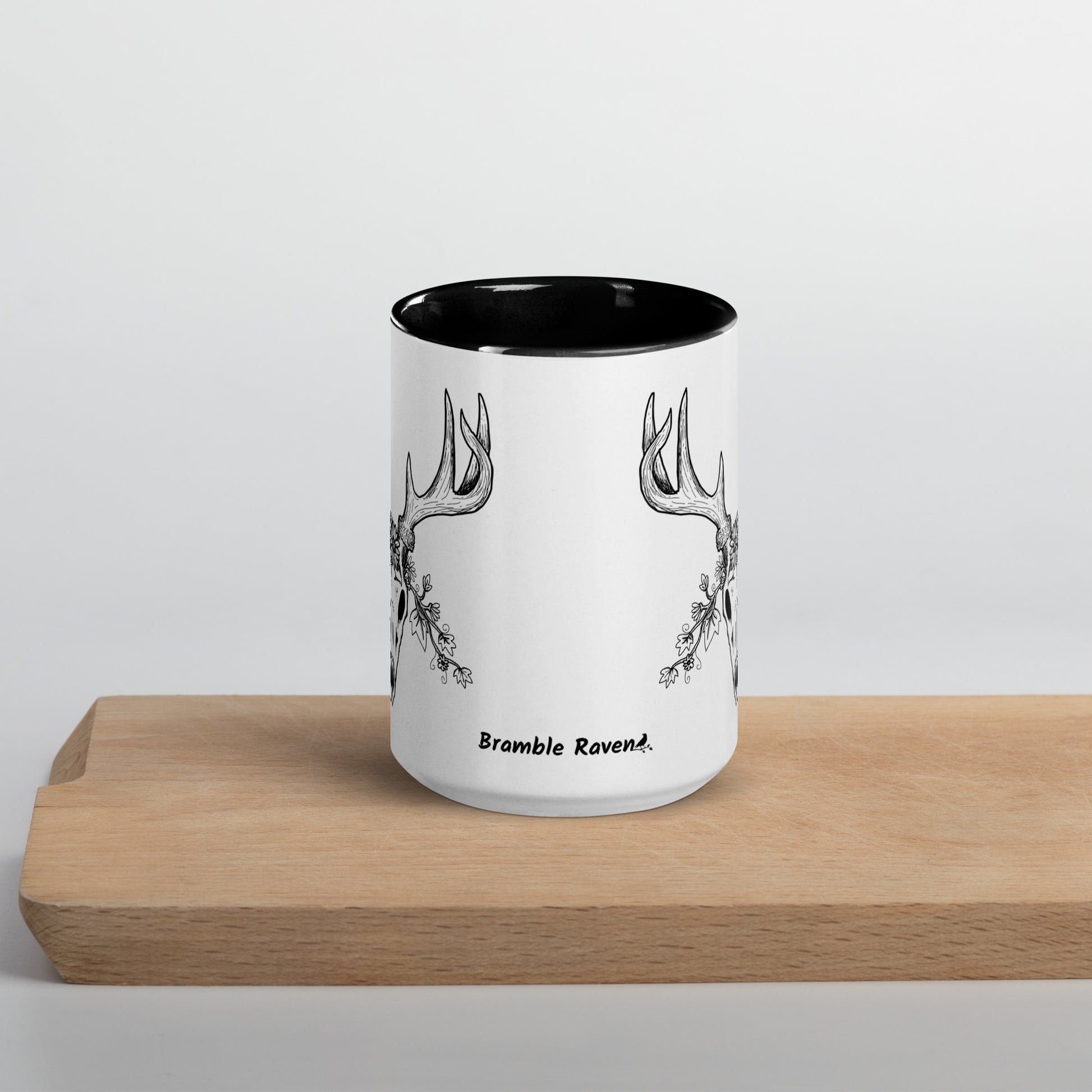 15 ounce ceramic mug with hand illustrated deer skull wreathed in flowers. Double-sided design. Mug has black rim, handle, and inside. Front view of mug shown sitting on wooden cutting board.